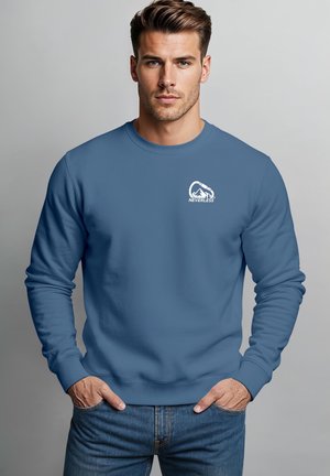 Mann trägt blauen Sweatshirt mit "NEVERLESS" Text und Berg-Logo auf der Brust, steht vor einem einfarbigen hellgrauen Hintergrund mit Händen in den Taschen.