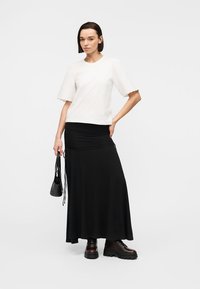 Γυναίκα που φοράει λευκή μπλούζα με φουσκωτά μανίκια, μακριά μαύρη φούστα, σκούρα μποτάκια με κορδόνια, κρατώντας μικρό μαύρο τσαντάκι μπροστά σε απλό φόντο.