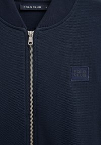 Navy blue textured jacket with silver zipper and stitched "POLO CLUB" logo patch on left chest.