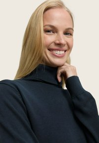 Pull en maille douce de couleur bleu marine foncé avec col roulé et manches longues. Présente un col haut et des poignets côtelés. Le modèle sourit doucement, les mains près du visage.