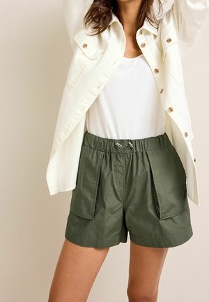 Mujer que lleva una camisa blanca con botones, top blanco y pantalones cortos verde oliva con bolsillos delanteros y cintura elástica con cordón.