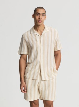 Giovane uomo che indossa una camicia a maniche corte a righe beige e bianche in maglia con bottoni e pantaloncini abbinati con coulisse, in piedi davanti a uno sfondo semplice.