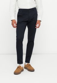 Pantaloni navy din bumbac, slim fit, cu buzunare laterale. Purtați cu pantofi din suede de culoare bronz și șosete albe până la gleznă, asortați cu o cămașă rayată.