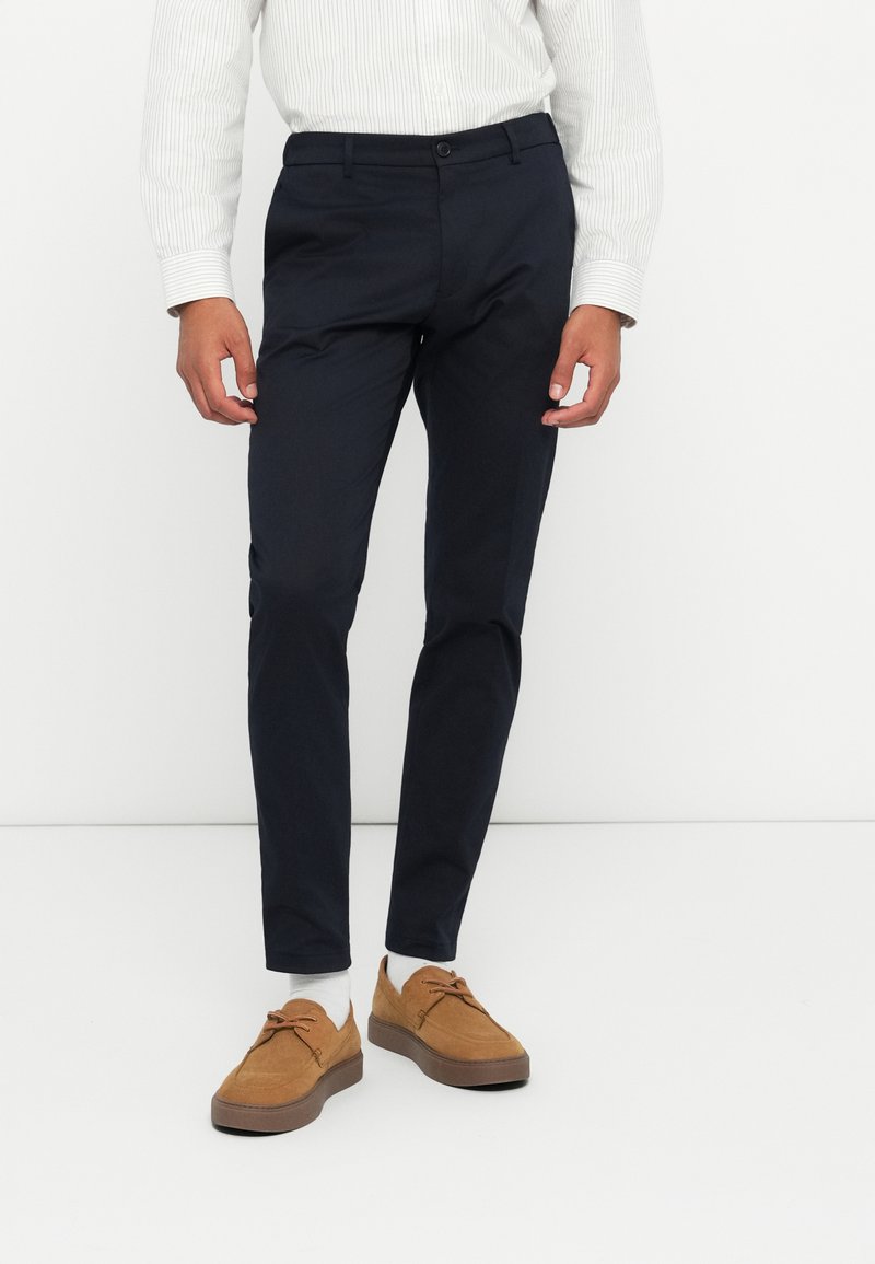 Pantaloni navy din bumbac, slim fit, cu buzunare laterale. Purtați cu pantofi din suede de culoare bronz și șosete albe până la gleznă, asortați cu o cămașă rayată.