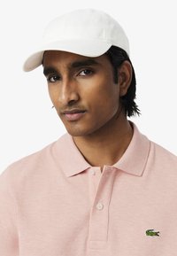 Vit basebollkeps och ljusrosa polo t-shirt med en texturerad yta och en grön Lacoste krokodil-logotyp på vänster bröst.