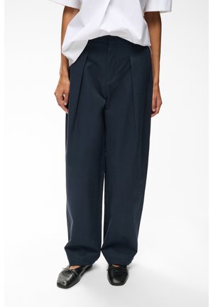 Personne portant un pantalon ample bleu marine foncé, des ballerines noires et une chemise blanche à manches courtes sur un fond blanc uni.