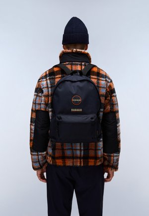 Laudan sininen reppu, jossa on pyöristetty muoto, etupuolella vetoketjullinen tasku ja logopatch. Käytetään kuvioidun oranssi-sinisen ruudullisen fleece-takin päällä.