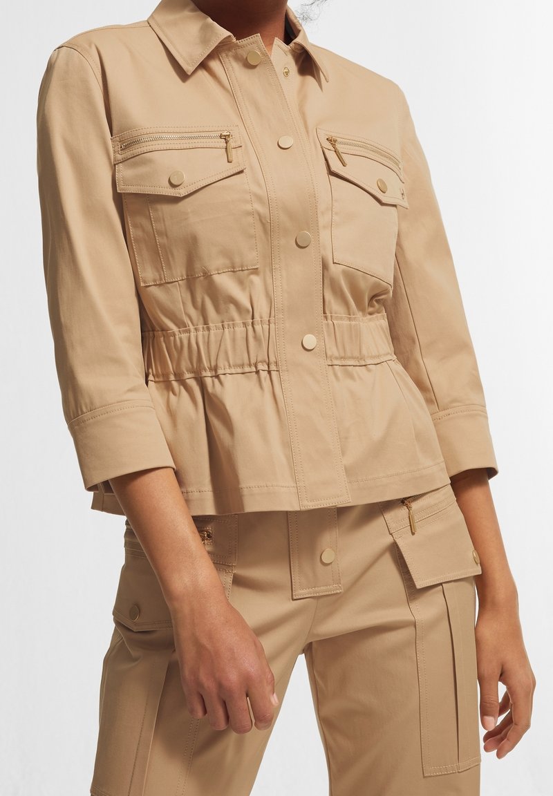 Beżowa kurtka użytkowa z dopasowaną talią, dwoma kieszeniami na piersi z zamkami, metalowymi akcentami i zapięciami na guziki. Teksturowany materiał, rękawy o długości trzech czwartych.