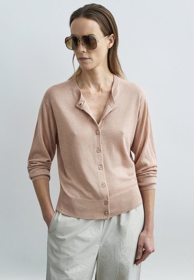Femme portant des lunettes de soleil marron surdimensionnées, un cardigan rose clair boutonné avec les manches retroussées, et un pantalon beige clair, debout avec une main dans la poche.