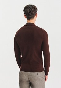 Brązowy sweter dziany o dopasowanym kroju i ściągaczach, pokazany z tyłu, w połączeniu z jasnoszarymi spodniami.