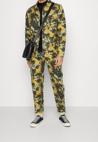 Kamouflagejacka och byxor i gröna, gula och blå toner; bomullstyg; har flera fickor; matchas med svarta sneakers och väska.