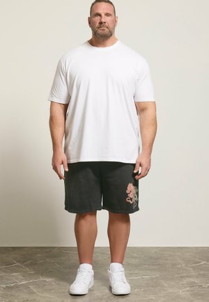 Erwachsener Mann stehend, trägt ein schlichtes weißes T-Shirt, schwarze Shorts mit Drachenmotiv, weiße Socken und weiße Turnschuhe vor neutralem Hintergrund.