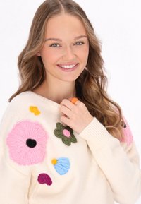 Crème gebreide trui met kleurrijke bloemenborduring in roze, groen, blauw en geel. Model draagt een oranje ring en roze oorbellen.