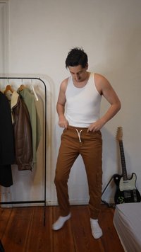 Άνδρας με λευκό φανελάκι και καφέ παντελόνι που ρυθμίζει τη ζώνη του σε δωμάτιο με κρεμάστρα ρούχων και κιθάρα πάνω σε ξύλινο πάτωμα.