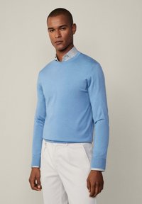Helles blaues Strickpullover mit Rundhalsausschnitt und langen Ärmeln, drüber getragen über einem collarierten, gestreiften Hemd, kombiniert mit weißen Hosen.