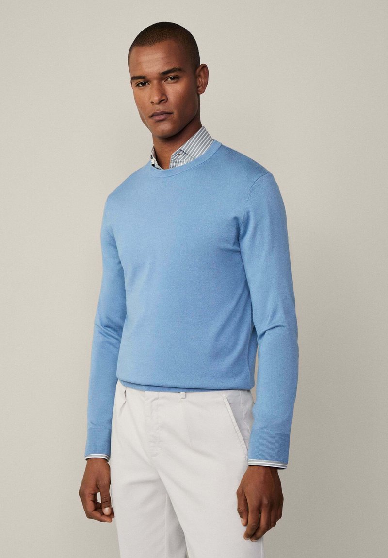 Helles blaues Strickpullover mit Rundhalsausschnitt und langen Ärmeln, drüber getragen über einem collarierten, gestreiften Hemd, kombiniert mit weißen Hosen.