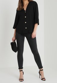 Svart skjorta med knappar och trekvartsärmar, prydd med guldknappar; kombinerad med mörka tajta jeans och svarta högklackade sandaler.