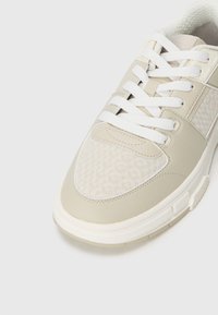 Beige athletic sneaker with textured leather and fabric panel featuring a logo pattern, white laces, and a thick white sole.