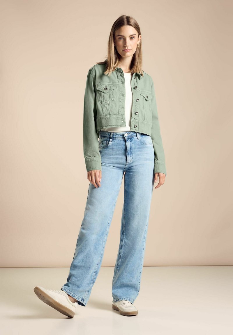 Veste en denim vert coupée avec deux poches poitrine, associée à un jean bleu clair à jambes larges et des baskets blanches sur un fond neutre.