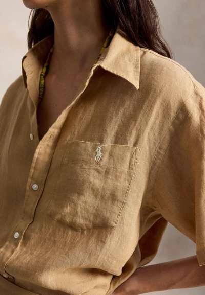 Moteris dėvi laisvą smėlio spalvos lino marškinius su kišene priekyje ir apykakle, papuoštus žaliai geltonu karoliukų vėriniu.