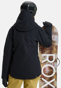 Black snowboard jacket with a hood, paired with yellow pants. The individual holds a snowboard featuring a nature-inspired design.