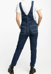 Jeanssalopette mit einer körperbetonten Passform, verstellbaren Trägern, zwei Gesäßtaschen und in einem dunkelblauen Waschton. Kombiniert mit schwarzen Stiefeln.