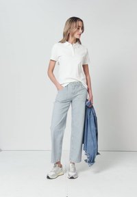 Femme debout, main dans la poche, portant un polo blanc, pantalon large rayé, baskets, tenant une veste en jean bleue en studio.