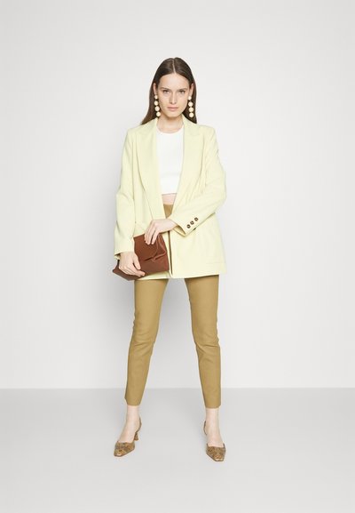 Κίτρινο σακάκι κομμένο πάνω από λευκό cropped τοπ, συνδυασμένο με καφέ fitted παντελόνι. Τα αξεσουάρ περιλαμβάνουν ένα καφέ clutch και παπούτσια με σχέδιο.