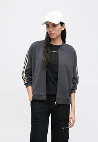 Ciemnoszara kurtka bomberka z zamkiem z przodu, ściągaczami na rękawach oraz beżowymi paskowymi akcentami na rękawach. Noszona na czarnej przylegającej koszulce i czarnych spodniach.