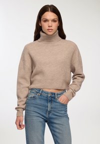 Jasno brązowy, krótki sweter z wysokim, prążkowanym kołnierzem, wykonany z miękkiego materiału; zestawiony z niebieskimi dżinsami z wysokim stanem.