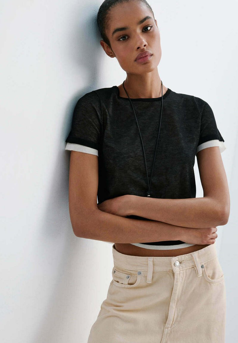 Jeune femme portant un crop top noir et un pantalon beige se tient les bras croisés contre un mur blanc, regardant la caméra.