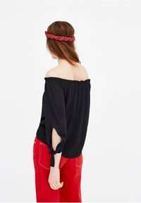 Schwarzes Off-Shoulder-Oberteil mit gerafftem Ausschnitt, Dreiviertelärmel mit Bindedetails, kombiniert mit roten weit geschnittenen Hosen.
