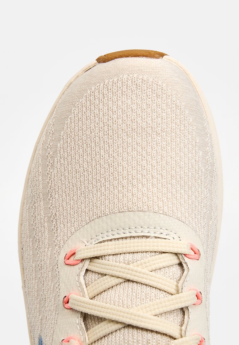 Zapatilla de color beige claro con parte superior de punto texturizado, acentos de cordones rosas y un tirador en el talón marrón. Presenta un diseño tradicional de cordones.