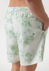 Zöld és fehér úszónadrág pálmafa mintával, rugalmas derékkal, oldalsó zsebekkel, valamint egy hátsó zsebbel, amely patentos záródással rendelkezik.