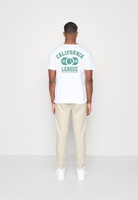 Vit t-shirt med grön tryckt text "CALIFORNIA LEAGUE 1774 COLLEGE CHAMPIONSHIP" på ryggen. Beige byxor och vita sneakers.