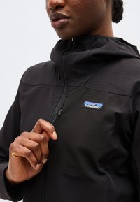 Zbliżenie na osobę ubraną w czarną kurtkę Patagonia, z częściowo rozpiętym zamkiem, ręką trzymającą suwak przy klatce piersiowej.