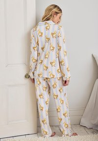 Nainen pukeutuneena valkoiseen pyjaman settiin, jossa on keltainen ankkakuvio, seisoo paljain jaloin valkoisen oven lähellä pehmeästi valaistussa huoneessa.