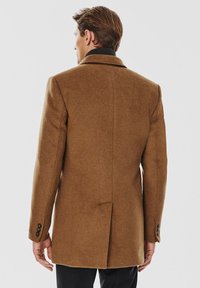 Brązowy wełniany płaszcz o dopasowanym kroju, z pojedynczym rozcięciem i mankietami zapinanymi na czarne guziki. Gładka tekstura i wyraźnie zarysowany projekt kołnierza.