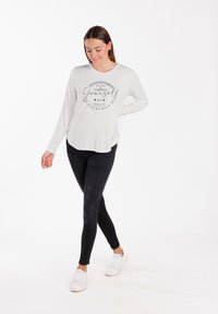 Langärmliges, hellgraues Oberteil mit schwarzem Drucktext, kombiniert mit schwarzen Skinny-Jeans und weißen Sneakern, vor einem weißen Hintergrund.