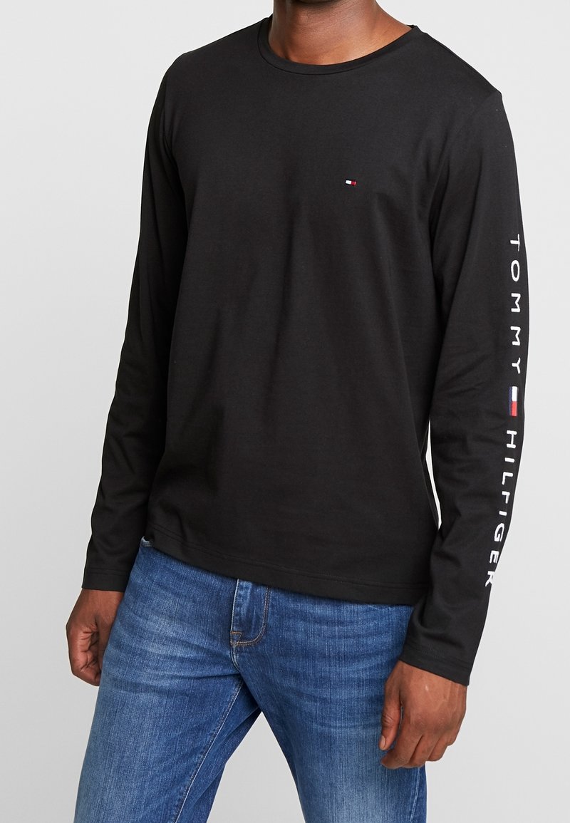 Schwarzes langärmliges T-Shirt aus Baumwolle. Verfügt über ein kleines Logo auf der Brust und "TOMMY HILFIGER" in Weiß entlang des Ärmels aufgedruckt.