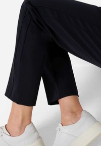 Pantalon noir raccourci avec une texture lisse, présentant une coupe droite et des coutures latérales discrètes, associé à des baskets blanches.