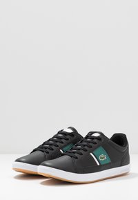Zapatillas negras hechas de material sintético texturizado, con acentos verdes, rayas blancas y una suela de goma. Diseño de cordones.