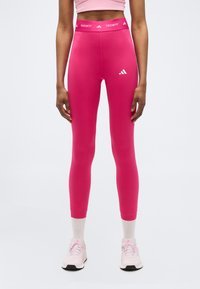 Γυναίκα που φοράει φωτεινά ροζ κολάν Adidas Techfit και ανοιχτό ροζ αθλητικά παπούτσια, στέκεται μπροστά σε ένα απλό ανοιχτό φόντο.