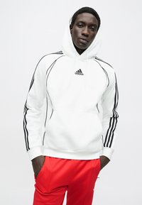 Bílá mikina s černými doplňky a třemi černými pruhy na rukávech, vybavená přední kapsou ve tvaru klokanky a kapucí se stahovací šňůrkou. Červené teplákové kalhoty.