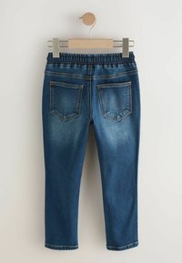 Pantaloni de denim albastri cu buzunare la spate și talie elastică atârnând pe un umeraș din lemn, cu fundal alb.