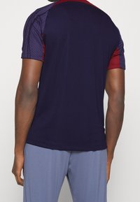 Camiseta atlética de color marino con mangas cortas; detalles en rojo en los hombros; tejido texturizado; acabado suave; combinada con pantalones de color azul claro.