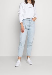 Jeans de denim azul claro con un corte relajado, combinados con una sudadera blanca con un logo negro. Zapatillas blancas completan el atuendo.