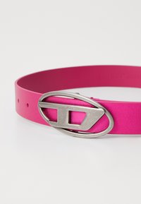 Rožinė guminė diržas su sidabriniu ovaliu užsegimu su išpjovomis, glotnia tekstūra ir keliais reguliavimo skylutėmis.