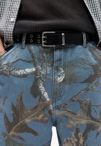 I pantaloni in denim presentano una base blu con un motivo di foglie e rami in tonalità terrose. Una cintura in pelle nera con accessori in argento completa il design.