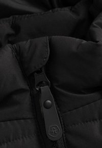 Juodas pūkų švarkas su storu užtrauktuku, lygiu paviršiumi ir siųtais audinių raštais, su apvalia apykaklės detalė.